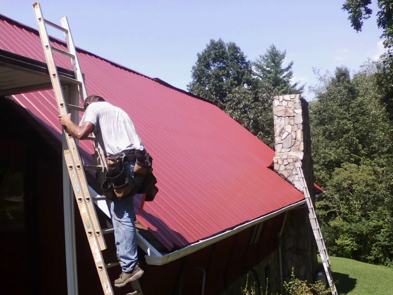 Roofer on a ladder beside a red metal roof and stone chimney — Soffit & Fascia Repair by Roofing Brothers in Christmas
